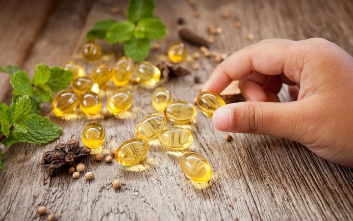 Botol kaca berisi minyak ikan berwarna keemasan dengan beberapa kapsul gel lembut di atas meja kayu, menunjukkan suplemen omega-3 yang populer